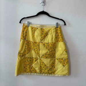 May include: A yellow patchwork skirt with a floral pattern. The skirt is hanging on a black hanger against a white wall. The skirt is made of quilted fabric and has a geometric design.