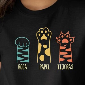 May include: Black t-shirt featuring three colorful cat paw designs with the words "Roca, Papel, Tijeras" underneath. The paw prints are blue, yellow, and orange. The shirt is a crew neck.