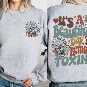 May include: Light gray sweatshirt with a floral kidney design on the front and back. The back of the sweatshirt has the text "It's A Beautiful Day To Remove Toxins" in a retro font. The sweatshirt is made of a soft material.