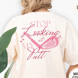May include: Ivory-colored t-shirt with a pink golf club graphic and the text "STOP Looking at MY Putt". The shirt has a relaxed fit and is made of a soft material. The text and graphic are centered on the back of the shirt.