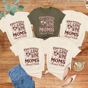 May include: Five t-shirts with the text "ORGANIZED GANG OF WINE MOMS Social Club" in a burgundy color. One shirt is olive green, and the others are natural. A pair of blue jeans, shoes, and a cup of coffee are also in the image.