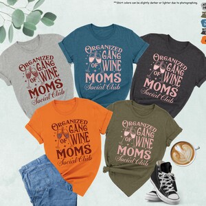 May include: A collection of t-shirts in various colors, including gray, teal, orange, dark gray, and olive green. Each shirt features the text "ORGANIZED GANG OF WINE MOMS Social Club" in a stylized font, with wine glasses. A pair of blue jeans and a black high-top sneaker are also visible.