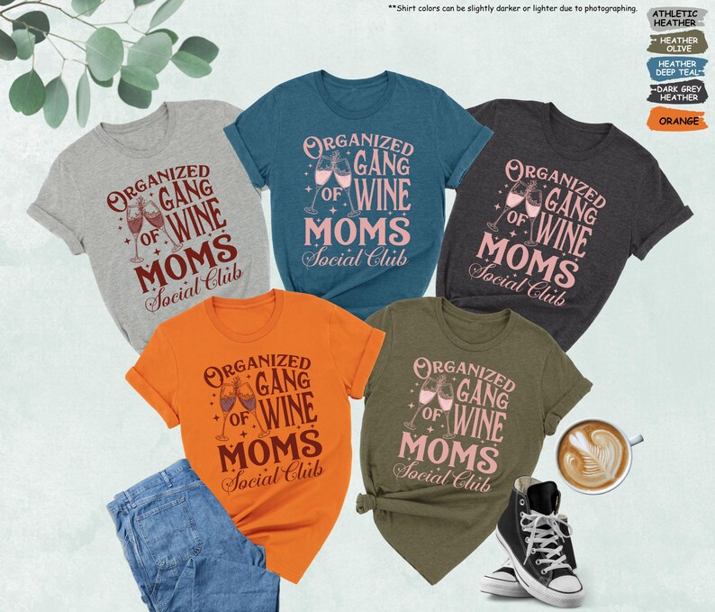 May include: A collection of t-shirts in various colors, including gray, teal, orange, dark gray, and olive green. Each shirt features the text "ORGANIZED GANG OF WINE MOMS Social Club" in a stylized font, with wine glasses. A pair of blue jeans and a black high-top sneaker are also visible.