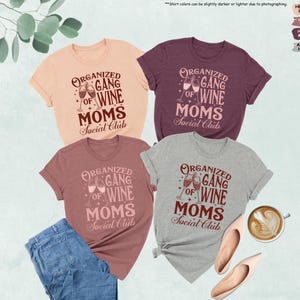 May include: Four t-shirts in peach, maroon, mauve, and athletic heather colors, each with the text "Organized Gang of Wine Moms Social Club" in a decorative font. The design includes wine glasses. A pair of jeans, shoes, and a cup of coffee are also in the image.