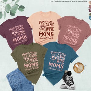 May include: A collection of t-shirts in various colors, including peach, mauve, maroon, olive, and deep teal. Each shirt features the text "ORGANIZED GANG OF WINE MOMS Social Club" in a stylized font, with wine glasses. A pair of jeans and sneakers are also visible.