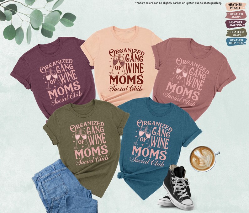 May include: A collection of t-shirts in various colors, including peach, mauve, maroon, olive, and deep teal. Each shirt features the text "ORGANIZED GANG OF WINE MOMS Social Club" in a stylized font, with wine glasses. A pair of jeans and sneakers are also visible.