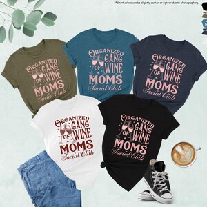 May include: Five t-shirts in various colors, including black, white, and olive green, are displayed. Each shirt features the text "Organized Gang of Wine Moms Social Club" in a pink and brown design, with wine glasses. A pair of jeans, a cup of coffee, and a pair of sneakers are also visible.