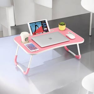 May include: A pink laptop desk with white legs and pink accents. The desk holds a laptop, tablet, smartphone, coffee cup, mouse, and a small potted plant. The desk is designed for use on a bed or sofa.