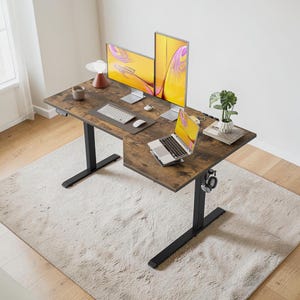 May include: A brown and black L-shaped standing desk with a wood-grain finish. The desk has two computer monitors, a laptop, keyboard, mouse, and a small potted plant. The desk is on a light-colored rug.