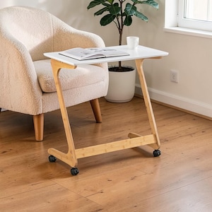 May include: A white, rectangular side table with a bamboo frame and rolling wheels. A book and a white mug sit on the table. The table is next to a beige armchair and a potted plant.