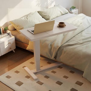 May include: A white adjustable laptop table with a laptop and a cup of coffee on top. The table is positioned over a bed with striped bedding. The table is supported by a white base on a patterned rug.