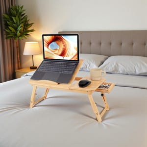 May include: A light-colored bamboo laptop desk with a built-in drawer and a side table. A laptop, a black mouse, and a beige mug are on the desk. The desk is on a bed with a white comforter.