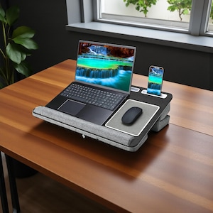 May include: A black laptop on a gray and black lap desk with a built-in mouse pad and phone holder. A black wireless mouse and a smartphone are also on the desk. The laptop screen displays a scenic waterfall.