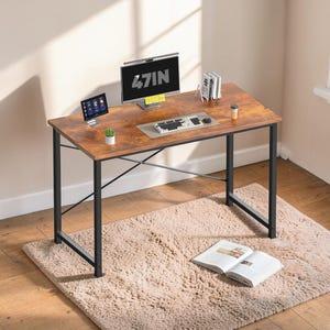 May include: A brown and black desk with a computer monitor displaying "47IN". The desk has a keyboard, mouse, and a small plant. A tablet and books are also on the desk. The desk is 47 inches wide.