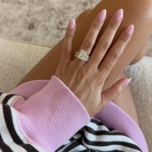 May include: A gold engagement ring set featuring a large rectangular radiant-cut center stone surrounded by a halo, paired with a matching eternity band encrusted with smaller rectangular stones, worn on a finger with light pink manicured nails.