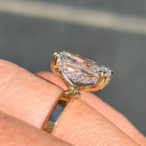 May include: A gold ring featuring a large, elongated rectangular-cut clear gemstone set in a prong setting. The gallery of the ring is accented with a row of small, round, clear pavé-set stones that follow the perimeter of the central stone.