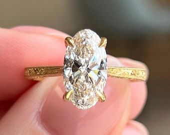 Könnte beinhalten: Ein Goldring mit einem großen, oval geschliffenen, klaren Edelstein, der von vier Krappen gehalten wird. Die Ringschiene besteht aus Gelbgold und weist auf der gesamten sichtbaren Außenfläche ein filigranes, graviertes Blumenmuster auf.