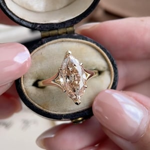 May include: A marquise-cut champagne-colored gemstone ring set in a gold band, presented inside a dark oval vintage jewelry box with the text 10 SANKEY ST., WARRINGTON. printed on the interior cream-colored fabric lining.