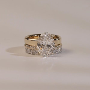 May include: A gold ring set consisting of a central oval-cut diamond solitaire ring, a thin gold band with a beaded texture, and a third band featuring a row of round-cut diamonds. The rings are stacked together to form a cohesive jewelry piece.
