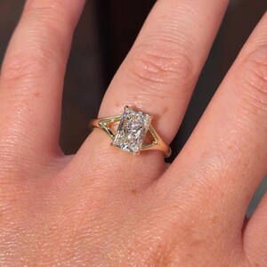 May include: A gold ring featuring a rectangular radiant-cut center stone held by four prongs. The band has a split-shank design that tapers toward the stone, creating a triangular opening on each side of the setting. The metal has a polished finish.