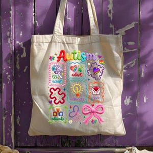 以下が含まれることがあります： ベージュのトートバッグに、カラフルな刺繍で「自閉症」の文字が施されています。バッグには、「受け入れる」、「適応する」、「提唱する」、「愛する」、「教育する」などの刺繍されたアイコンと単語が描かれています。ピンクのリボンもバッグに刺繍されています。