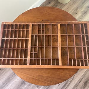 May include: A vintage wooden printer's tray with multiple small compartments. The tray is rectangular and has a warm, brown color. It is resting on a round wooden table, showcasing its design and potential use for organizing small items.