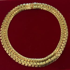 May include: A gold chain necklace with a woven design. The necklace is on a red velvet background.