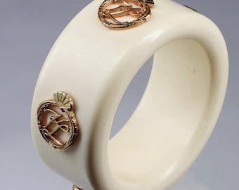 May include: White bangle bracelet with gold accents. The bracelet features two gold emblems with a stylized letter "L" inside a circle.