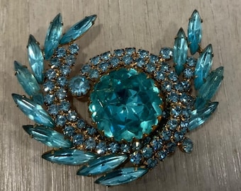 May include: Vintage brooch with a central, large, faceted, teal-colored stone surrounded by smaller, round, teal stones. Teal marquise-shaped stones form a wreath around the central design. The metal setting is gold-toned.