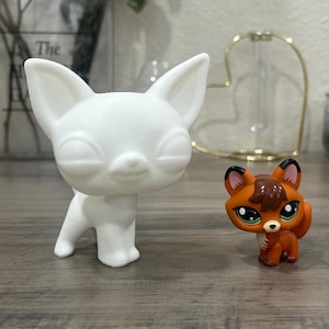 May include: Two small animal figurines. One is a white, stylized dog with closed eyes. The other is a small orange fox with brown and black accents and large green eyes. Both are standing on a wooden surface.