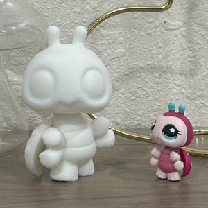 May include: Two small figurines. One is a large, white, cartoon-like insect with large eyes and a segmented body. The other is a smaller, pink and red insect figurine with large eyes and a shell-like back. Both are on a wooden surface.