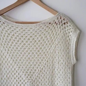 May include: Off-white, sleeveless crochet top with a scoop neck. The top is made of a loosely woven, open stitch pattern. The garment is hanging on a wooden hanger against a white background. The crochet design is visible.
