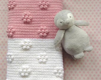 Puede incluir: Una manta de bebé hecha a mano en rosa y blanco, con un diseño floral. La mitad superior es rosa con detalles de flores en relieve, mientras que la mitad inferior es blanca. Un juguete de peluche de pingüino gris descansa junto a la manta.