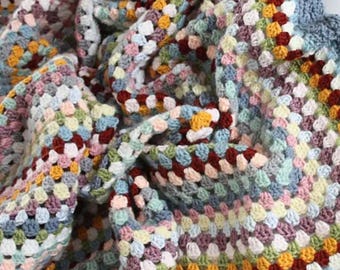 May include: A colorful, handmade crochet blanket. The blanket features a granny square design with a variety of colors including blue, red, yellow, and pink. The blanket is folded, showing the texture and pattern of the yarn.