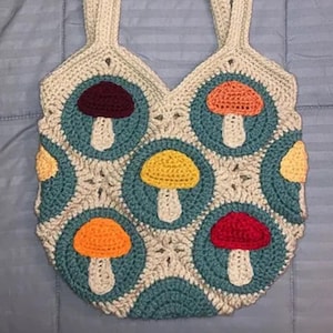 May include: A cream-colored crochet tote bag featuring a pattern of colorful mushrooms. The mushrooms have red, orange, and yellow caps with white stems, set against teal circles. The bag has two cream-colored straps.