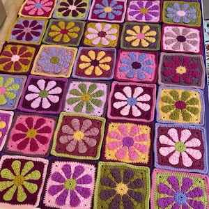 May include: A colorful, handmade crochet blanket featuring a patchwork design of square blocks. Each square showcases a vibrant flower motif in shades of pink, purple, yellow, and green. The blanket is a cozy, decorative item.