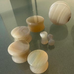 May include: A collection of various sizes of ear plugs made from a light-colored, swirled stone. The plugs are in shades of white, cream, and tan, with visible banding patterns. Some are rounded, while others are cylindrical.