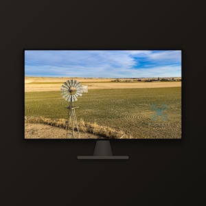 May include: A computer monitor displaying a landscape photograph of a metal windmill standing in a green field under a blue sky with light clouds. The screen shows a rural scene with dry grass and a small watermark logo in the bottom right corner.