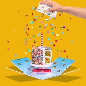 May include: A pop-up birthday cake box with a single lit candle on top. The box is open and confetti is falling out. The cake is decorated with colorful sprinkles and has a white frosting.