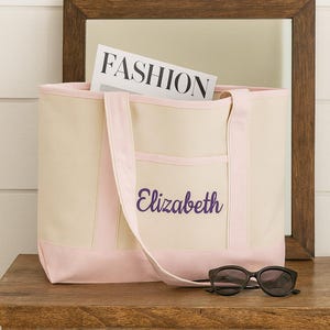 May include: A canvas tote bag with pink accents and the name "Elizabeth" embroidered in purple. The bag contains a magazine with the word "FASHION" on the cover. Black sunglasses rest on the wooden surface.