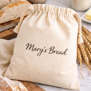 May include: A beige drawstring bread bag with the text "Mary's Bread" in a script font. The bag is filled with bread, and there are slices of bread on a wooden cutting board. Wheat stalks and a jar of flour are also visible.