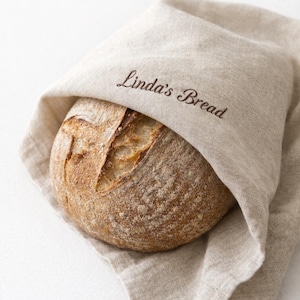 May include: A round loaf of bread, partially wrapped in a beige linen cloth. The bread has a golden-brown crust with a scored top. The cloth is embroidered with the words "Linda's Bread" in a brown script.