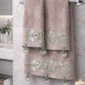 May include: Two mauve bath towels hanging on a chrome towel rack. The towels have an embroidered floral design in white and green along the bottom edge. Decorative tassels hang from the embroidered border. The towels are in a bathroom setting.