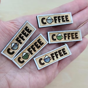 May include: A collection of rectangular wooden coffee-themed embellishments. Each piece features the word "COFFEE" in a bold, dark font, along with a small coffee cup illustration. The cups are colored in shades of blue, green, and white.