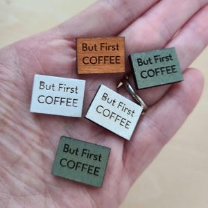 May include: A collection of small, square wooden tags with the phrase "But First COFFEE" engraved on them. The tags are in various colors, including brown, white, and green. The image is a close-up shot.