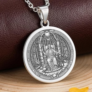 May include: A silver pendant necklace featuring a detailed circular medallion with an engraved design. The pendant depicts a seated figure within a detailed scene, suspended from a delicate chain. The necklace is displayed on a brown surface.