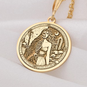 Op de afbeelding: Een goudkleurige ketting met een ronde hanger. De hanger toont een gedetailleerde afbeelding van een vrouw, half skelet, half levend, omringd door schedels en tekst. De ketting is ook goudkleurig.