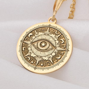 Può includere: Una collana con pendente dorato con un pendente circolare inciso con un disegno a occhio. Il pendente è sospeso a una catena sottile. Il disegno dell'occhio è circondato da elementi celesti, tra cui stelle e lune crescenti. La collana è presentata su tessuto bianco.