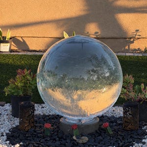 Puede incluir: Una fuente esférica de vidrio transparente sobre una base de hormigón, rodeada de piedras negras y blancas, suculentas y faroles decorativos. La imagen está tomada al aire libre, con el texto "Fountain & Flora" en la parte inferior.