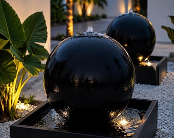 Könnte beinhalten: Zwei schwarze, kugelförmige Wasserbrunnen, die jeweils in einem quadratischen schwarzen Becken stehen. Wasser fließt von der Spitze jeder Kugel und erzeugt Wellen im Becken. Die Brunnen stehen im Freien und werden in der Dämmerung beleuchtet.